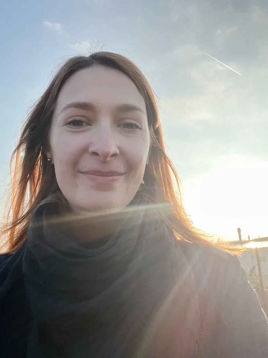 Selfie blondýny v tmavém šálu proti protislní na obloze s letadlovou contrail.