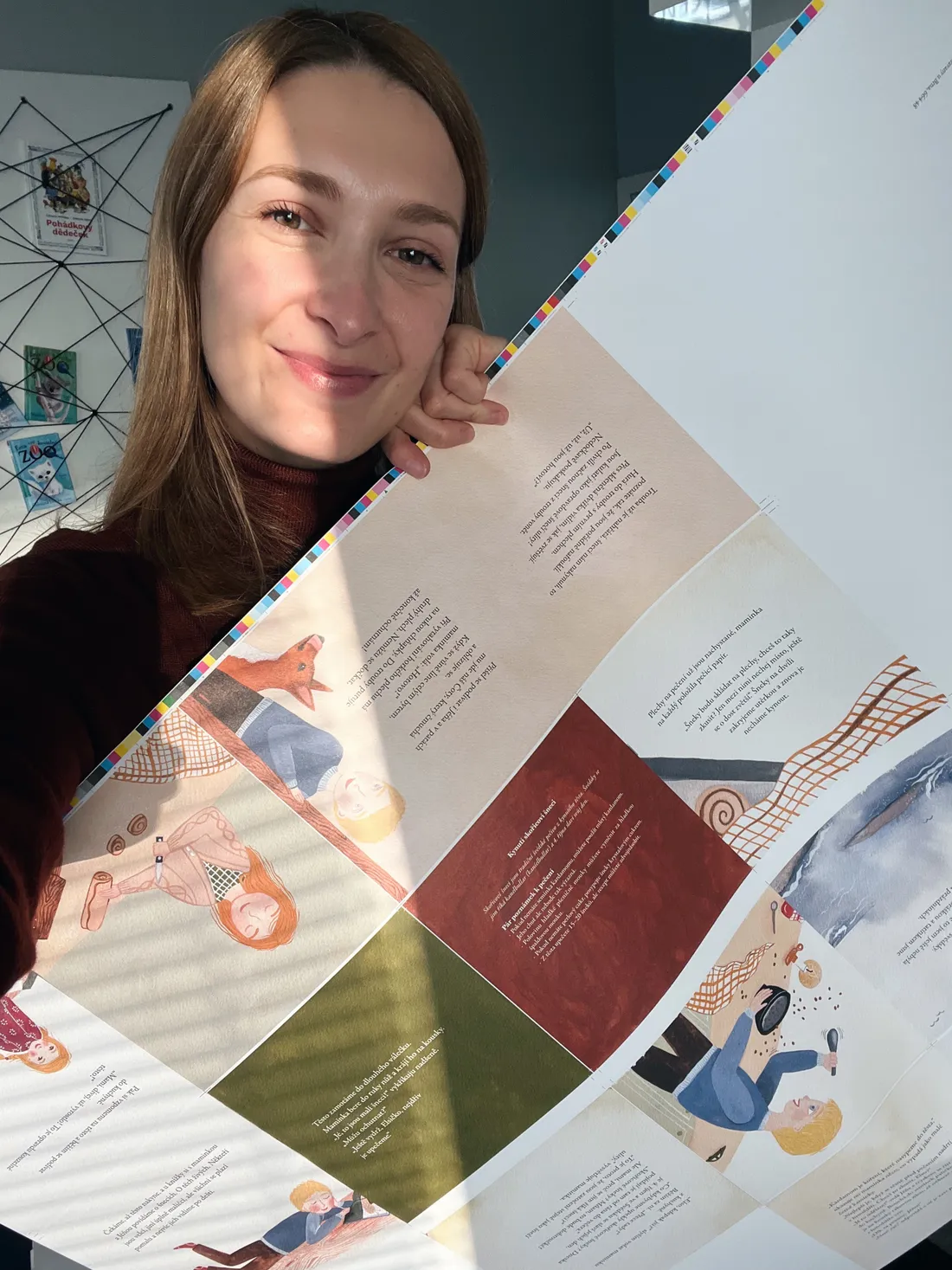 Usmívající se žena drží velký tiskový arch s ilustracemi a textem dětské knihy.