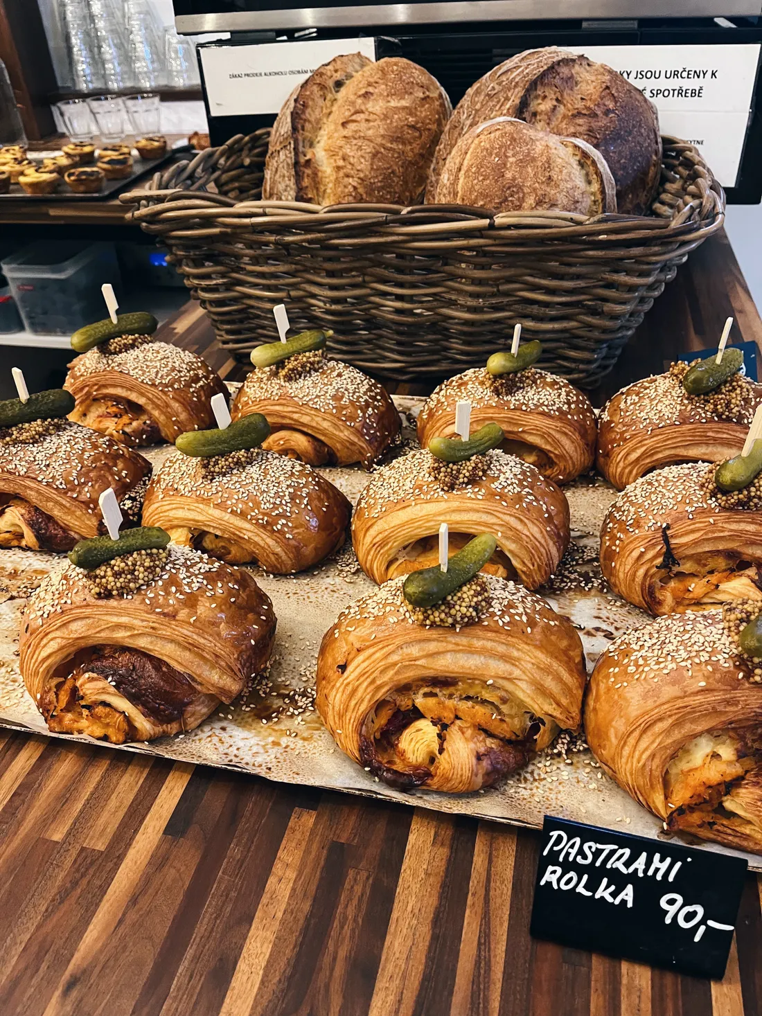 Slané rolky z listového těsta s pastrami a sezamem, hořčicí a okurkou na pultu, v pozadí proutěný koš s chlebem.