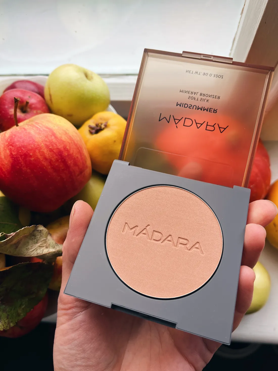 Ruka drží otevřený bronzer Mádara Midsummer před mísou s jablky na parapetu.