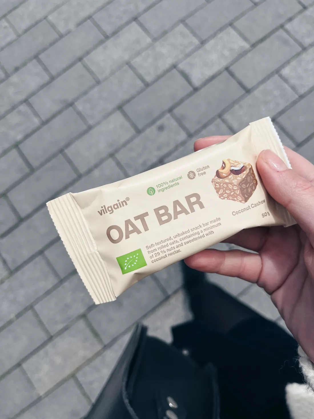 Ruka drží ovesnou tyčinku Vilgain Coconut Cashew na šedé dlažbě