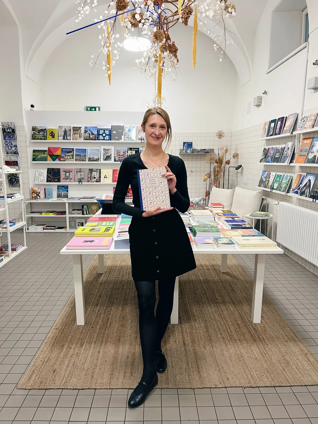 Autorka v černých šatech drží kuchařku před stolem plným knih v Book Therapy