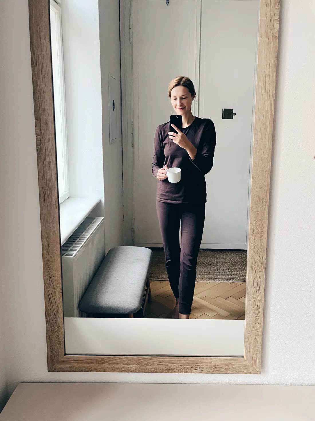 Selfie ve zrcadle: žena v tmavě hnědém domácím kompletu drží telefon a bílý hrnek v jasném pokoji.