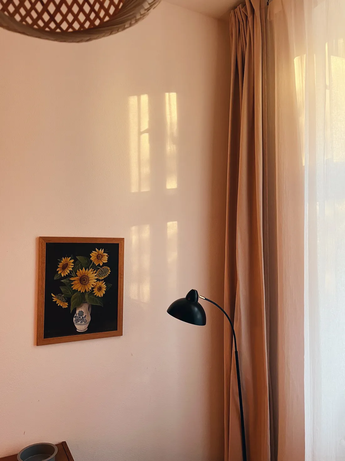 Na broskvové stěně visí obraz slunečnic, vedle černá lampa a světlo okna na závěsu.