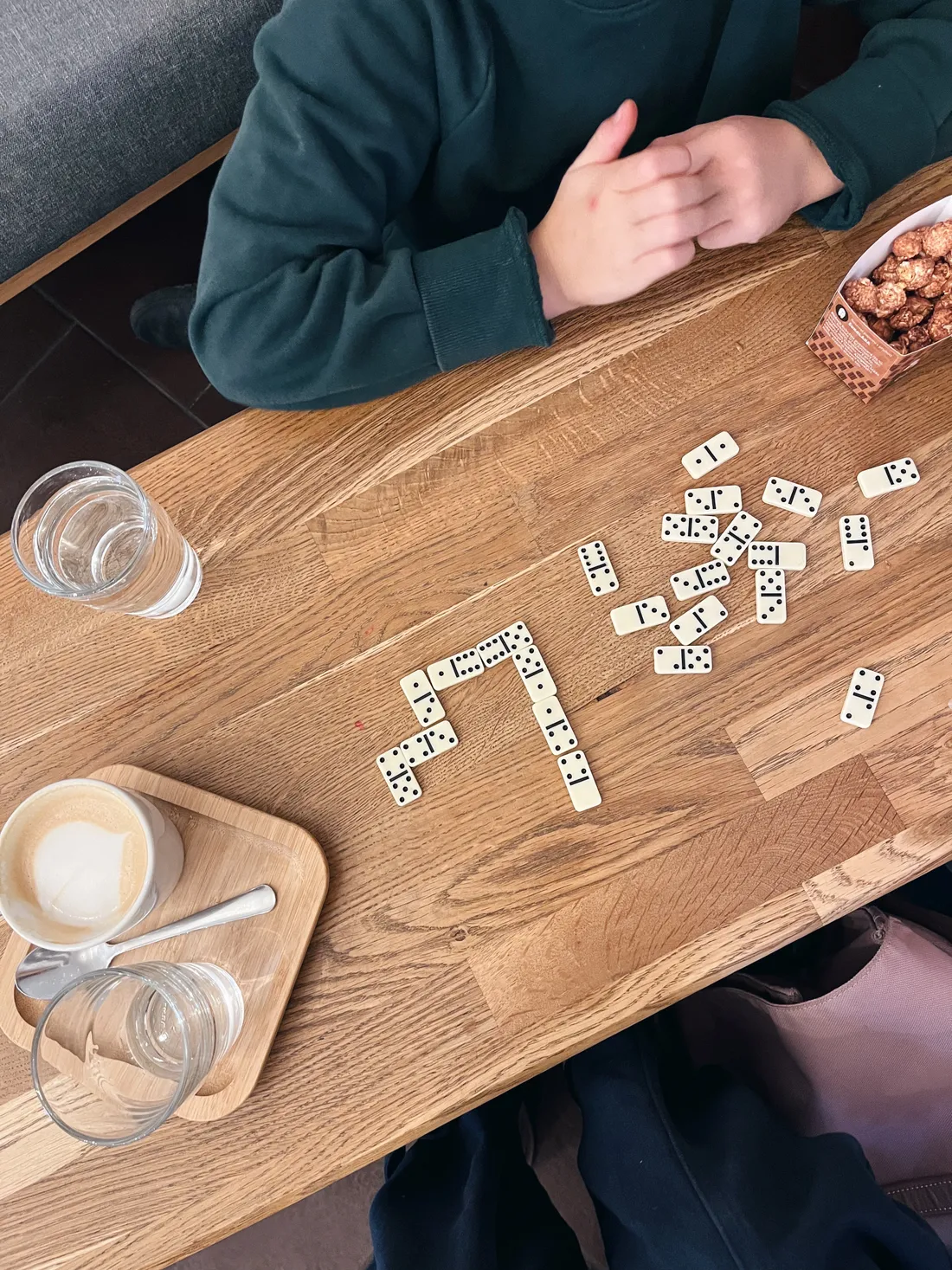 Domino na dřevěném stole, káva s latte art, sklenice vody a krabička popcornu u osoby v zelené mikině.