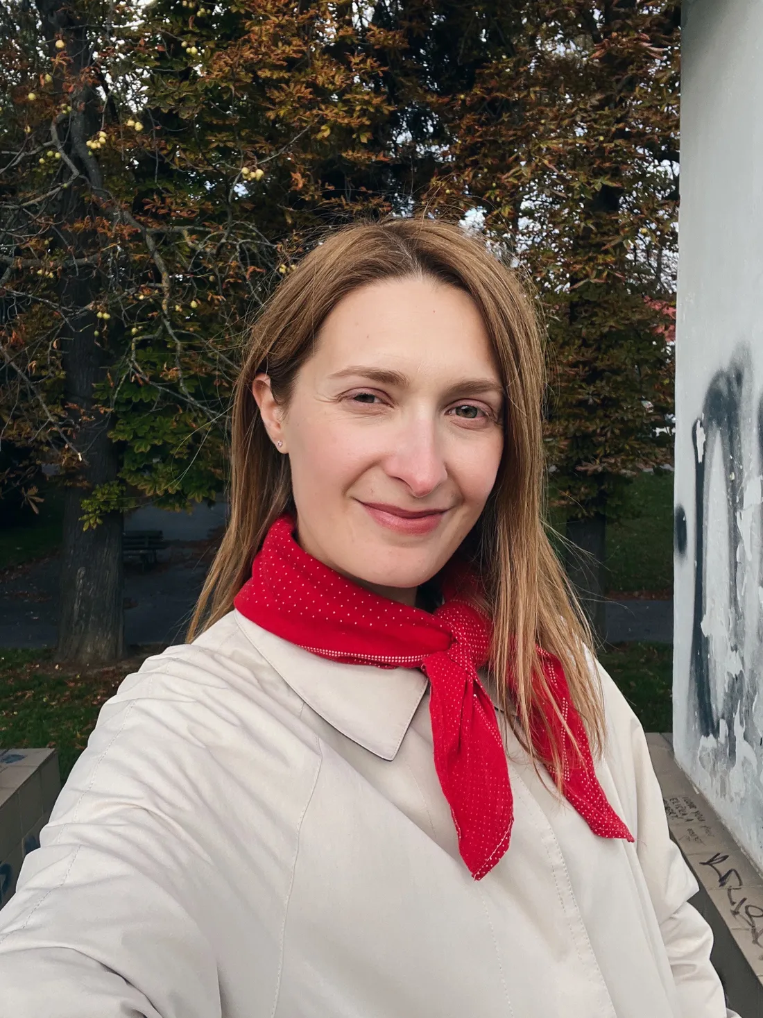 Žena v béžovém kabátu s červeným šátkem s puntíky pod podzimními stromy.