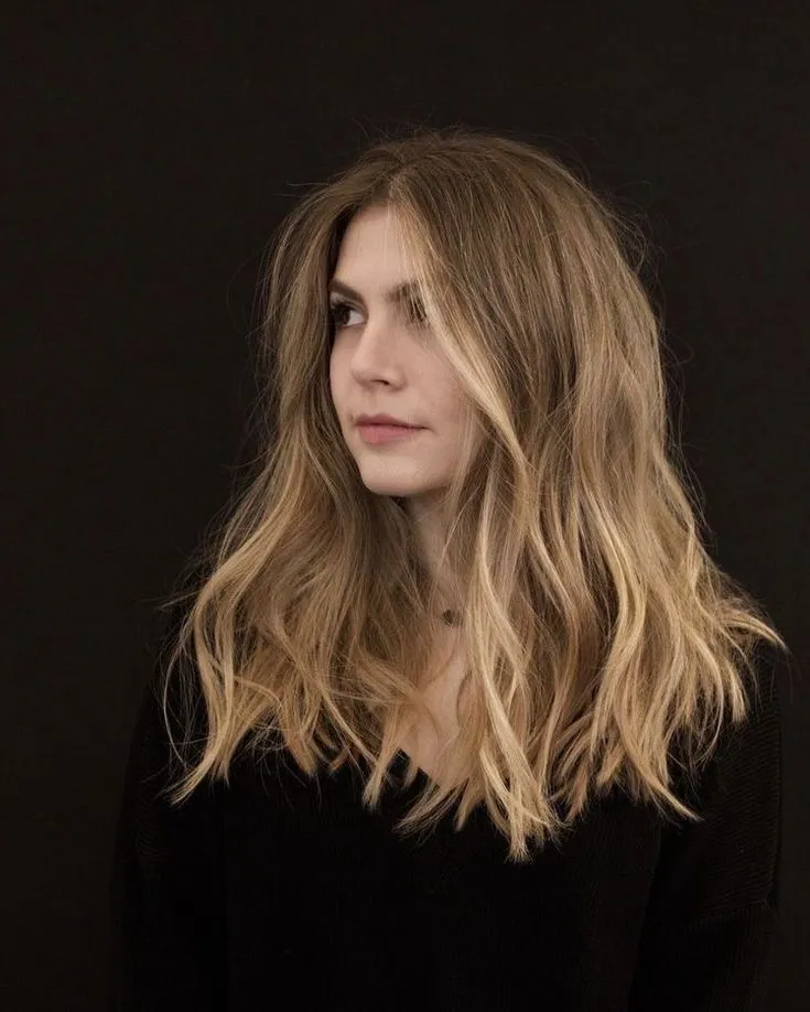 Mladá žena s dlouhými vlnitými blond vlasy v melíru, hledící stranou na černém pozadí.