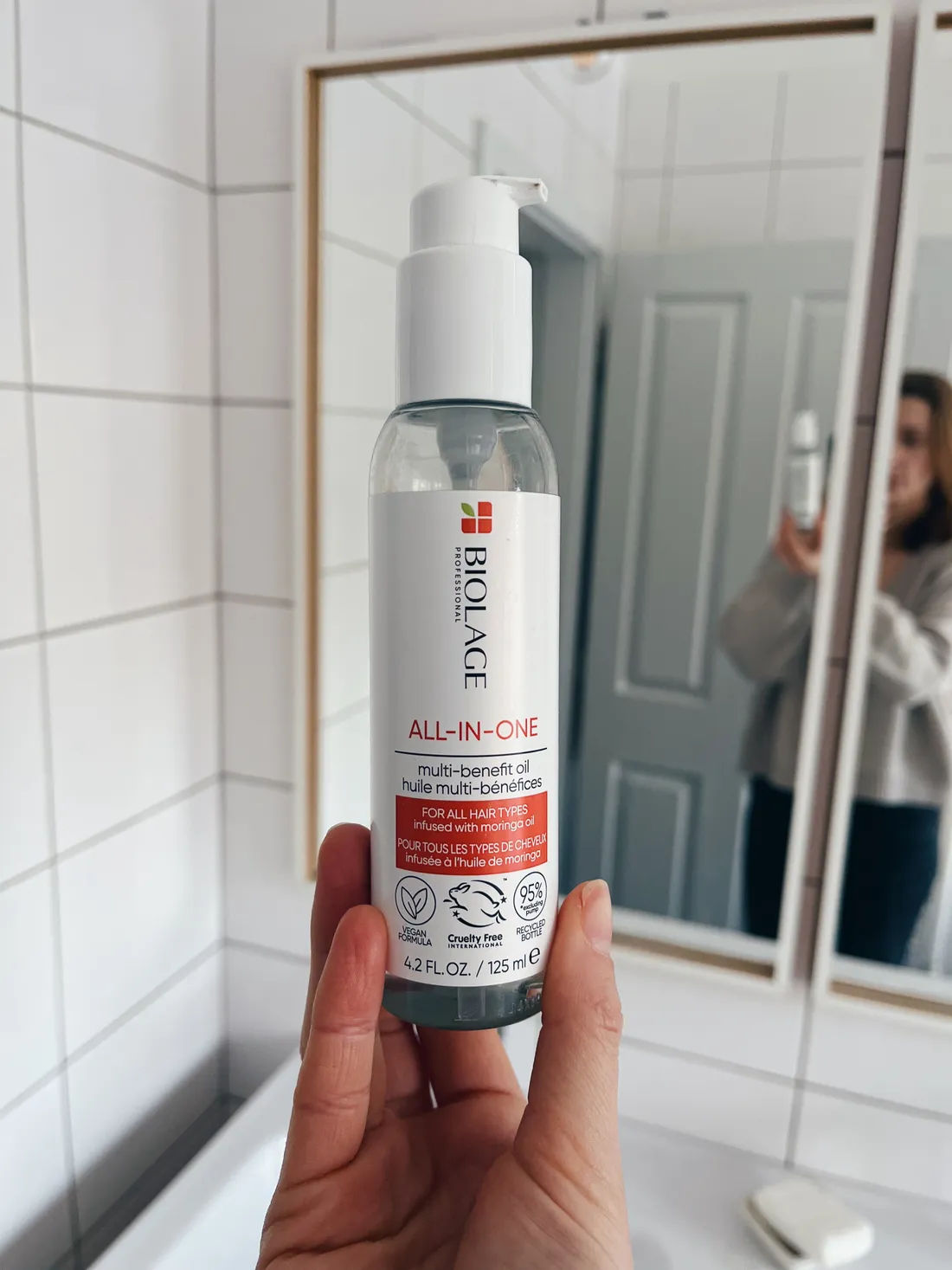 Ruka drží lahvičku Biolage All-in-One oleje v koupelně s bílými obklady.
