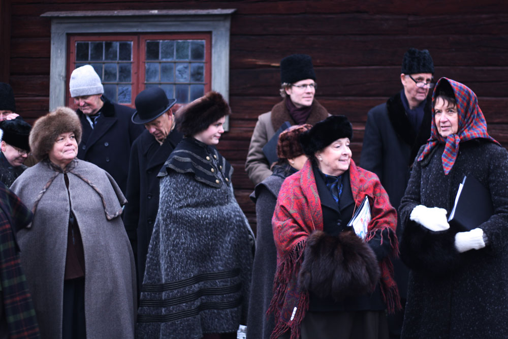 Skupina lidí v historických zimních oděvech s kožešinovými čepicemi a plášti před tmavou dřevěnou stěnou.