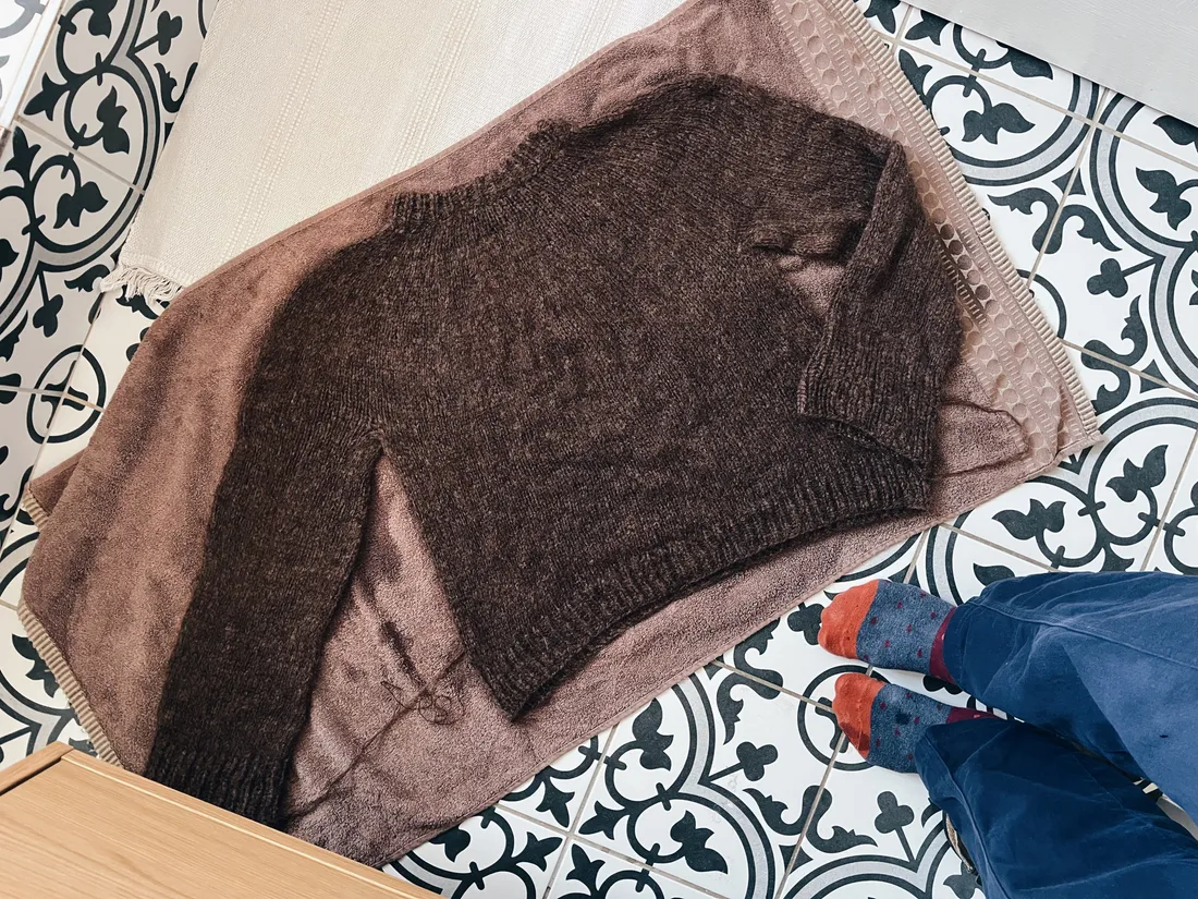 Tmavě hnědý svetr rozložený na hnědém ručníku na černobílé kachličkové podlaze při sušení.