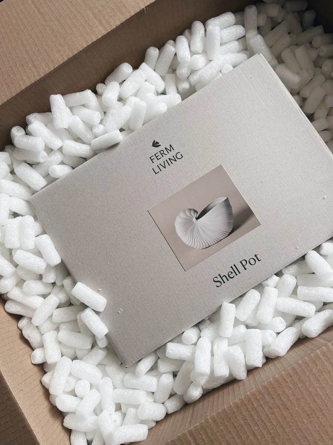 Šedá krabice Shell Pot od Ferm Living v kartonu vyplněném bílými výplňovými kuličkami.