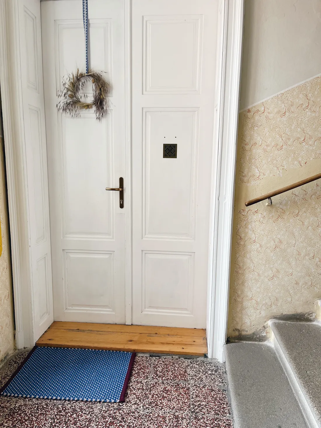 Bílé dveře se sušeným věncem, modrou rohožkou a schodištěm se zábradlím.