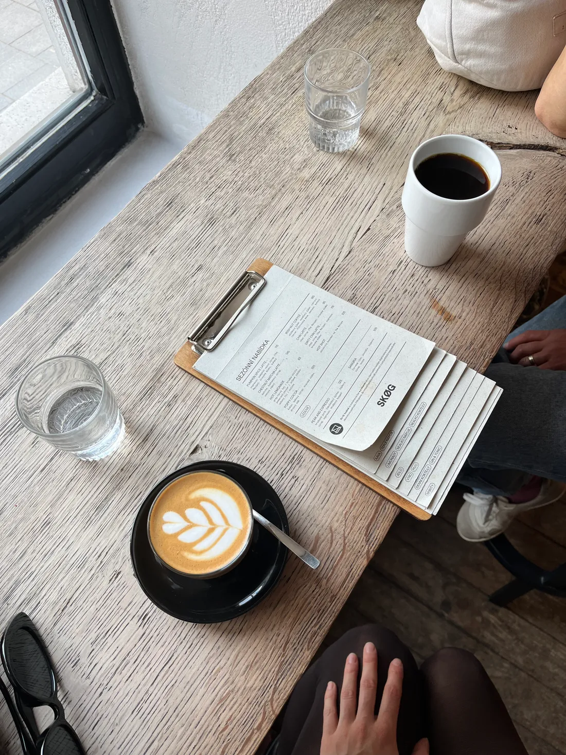 Dřevěný stůl v kavárně s cappuccinem, bílým hrnkem kávy, sklenkami vody a menu SKØG na dřevěné desce.