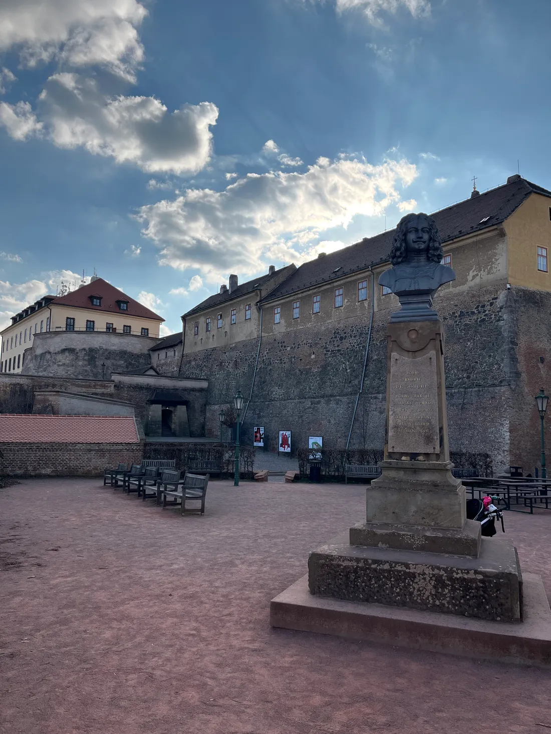 Bronzová busta na kamenném podstavci na nádvoří hradu Špilberk se slunečními paprsky na obloze.