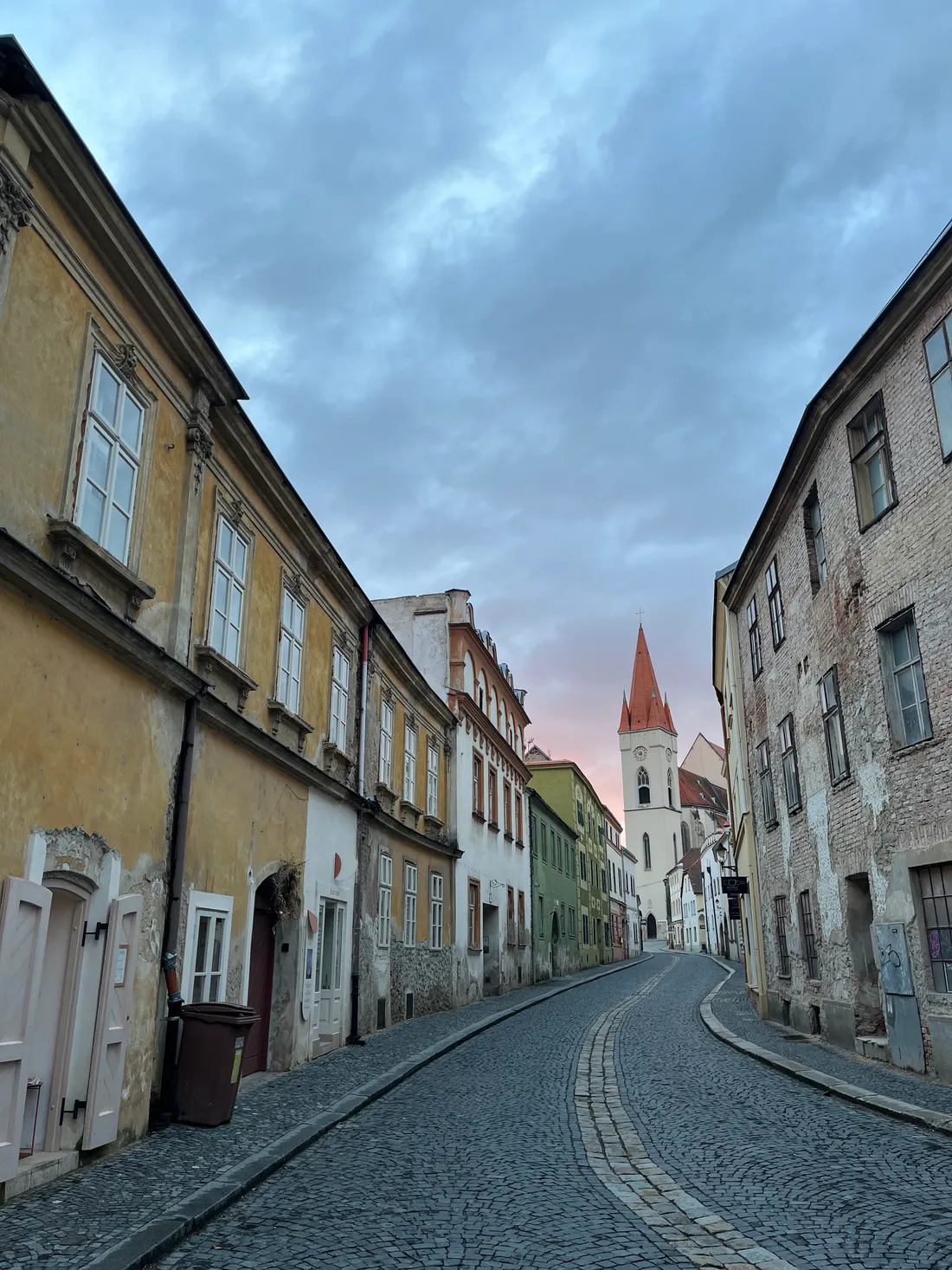 Úzká dlážděná ulička mezi historickými domy k bílé kostelní věži s červenou špičkou za soumraku.