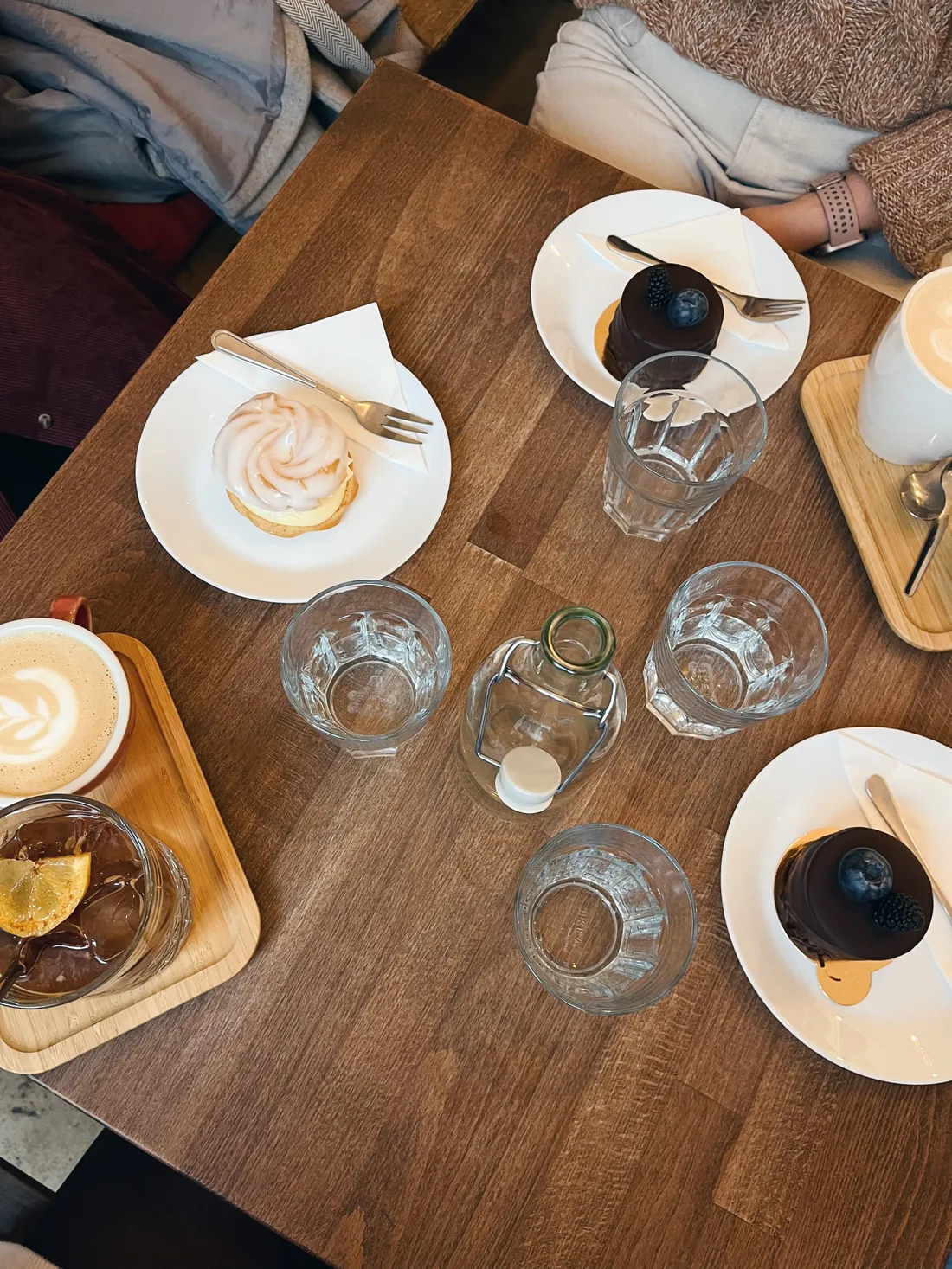 Dřevěný stůl s čokoládovými dortíky, větrníkem, kávou s latte art a lahví vody v kavárně.