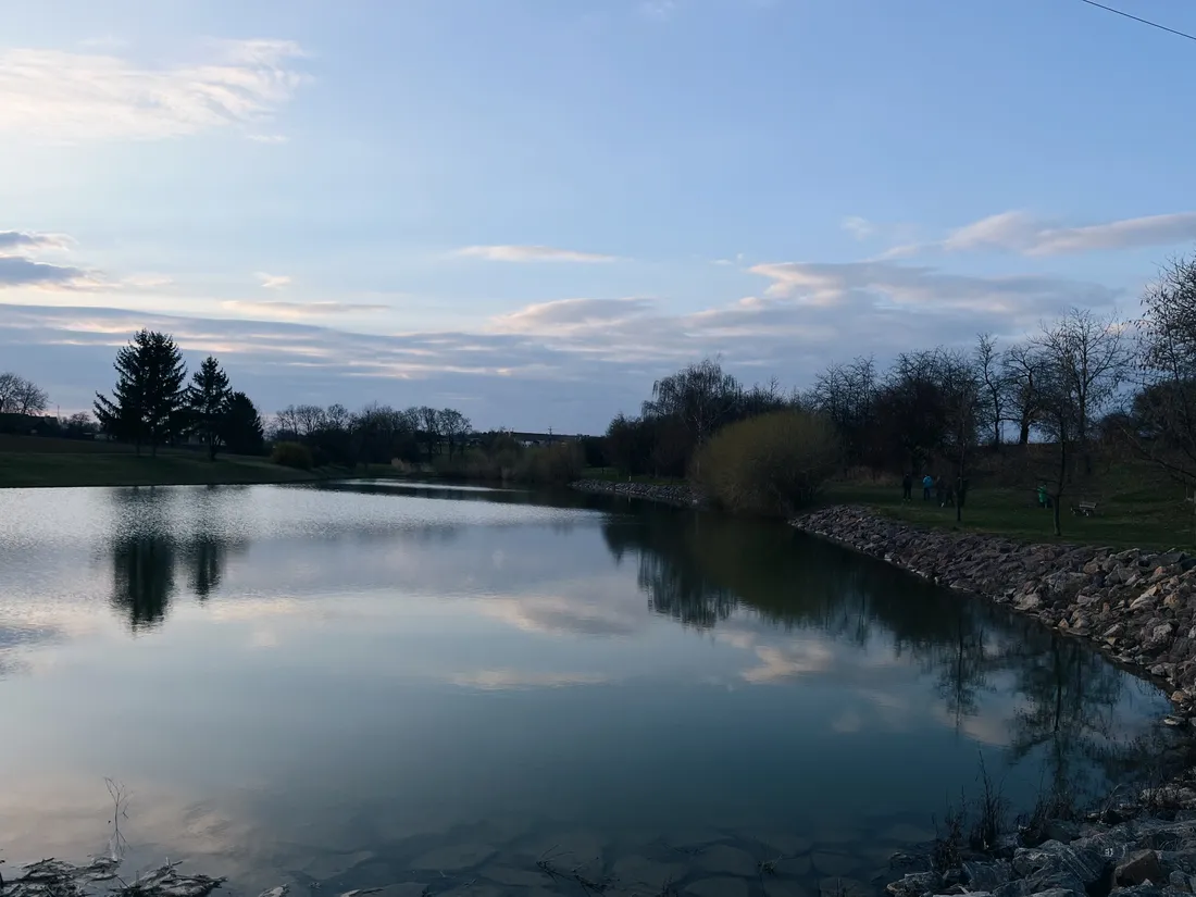 Klidná hladina rybníka odráží nebe s mraky, jehličnany na břehu a kamenné opevnění napravo.