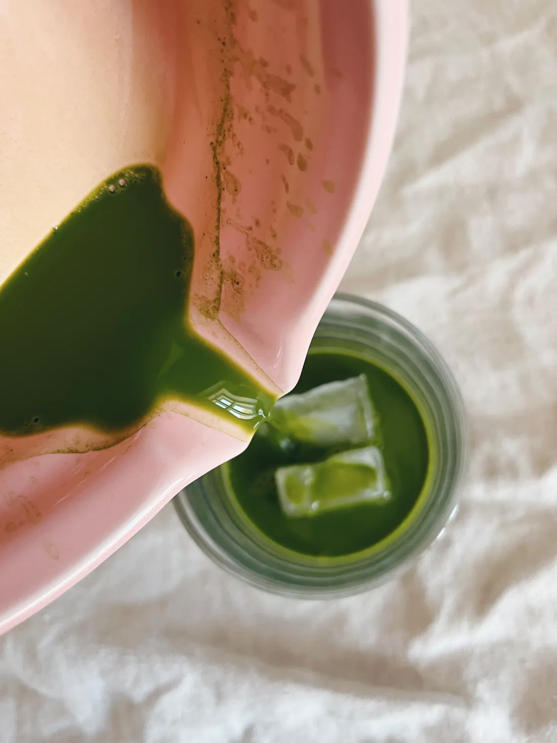 Zelený matcha čaj se nalévá z růžové misky do sklenice s ledovými kostkami na bílém ubrusu.