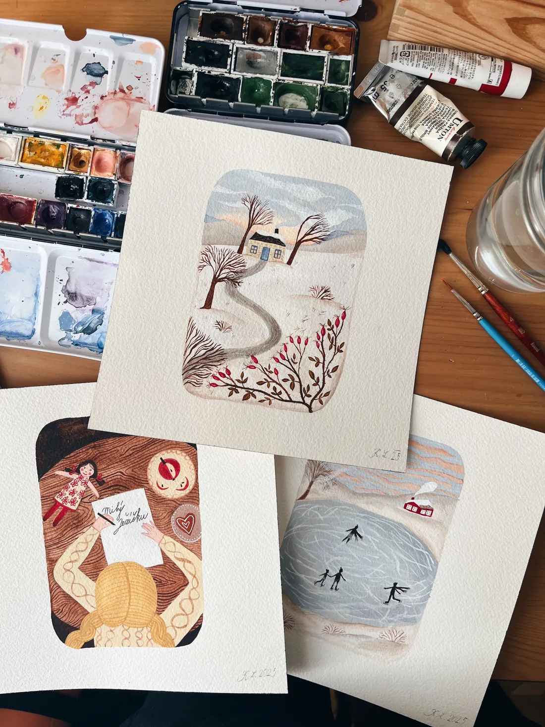 Dřevěný stůl s akvarelovými ilustracemi, paletou, štětci a sklenicí vody při malování.