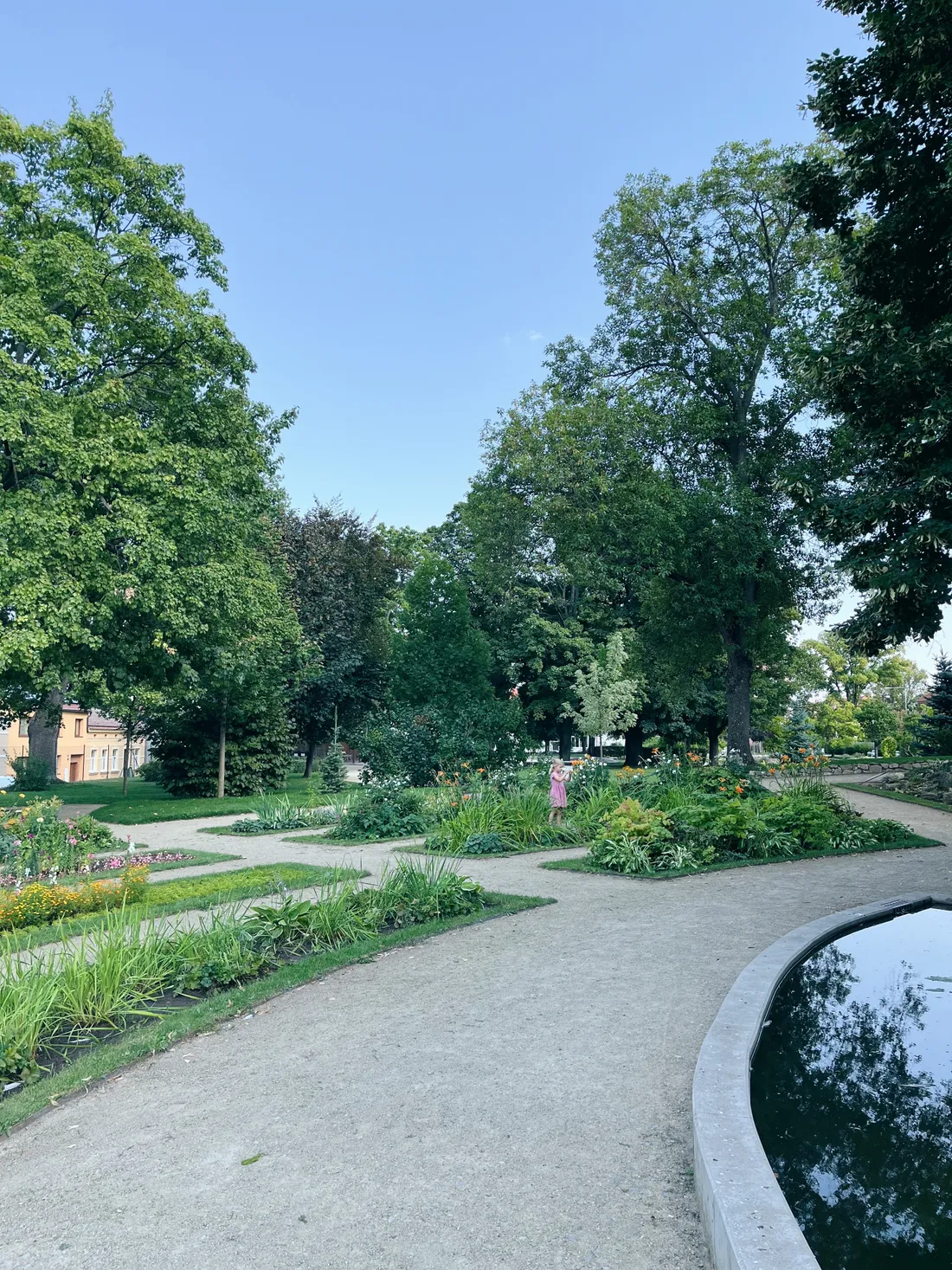 Park se štěrkovými cestami, květinovými záhony a stromy, v dálce postava v růžových šatech.