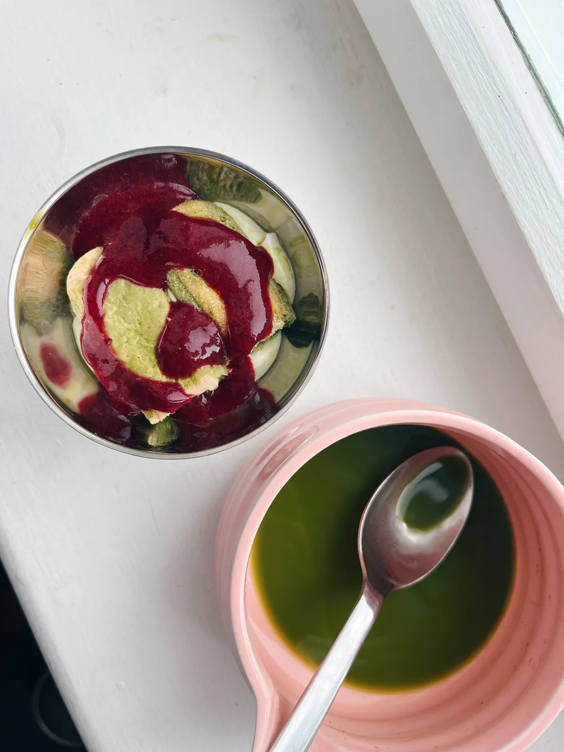 Růžový hrnek matcha a miska dezertu se zeleným piškotem a červenou omáčkou na bílém parapetu.