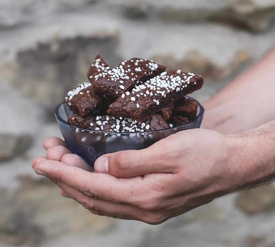 Ruce drží modrou skleněnou misku plnou tmavých čokoládových sušenek chokladsnittar posypaných perlovým cukrem na kamenitém povrchu.