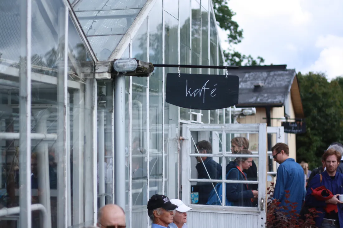 Černá cedule kafé visí na bílém skleníku zimní zahrady s lidmi u vchodu.