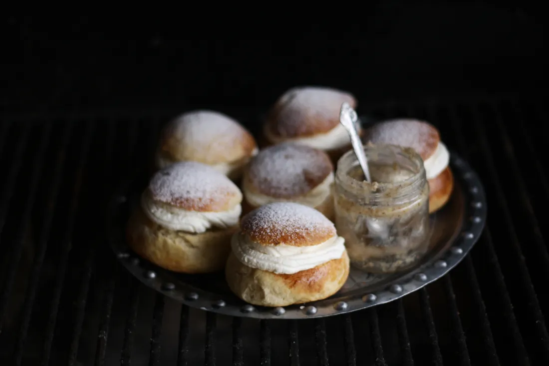 Šest semla bochánků se šlehačkou a sklenice mandlové pasty se lžičkou na kovovém talíři.