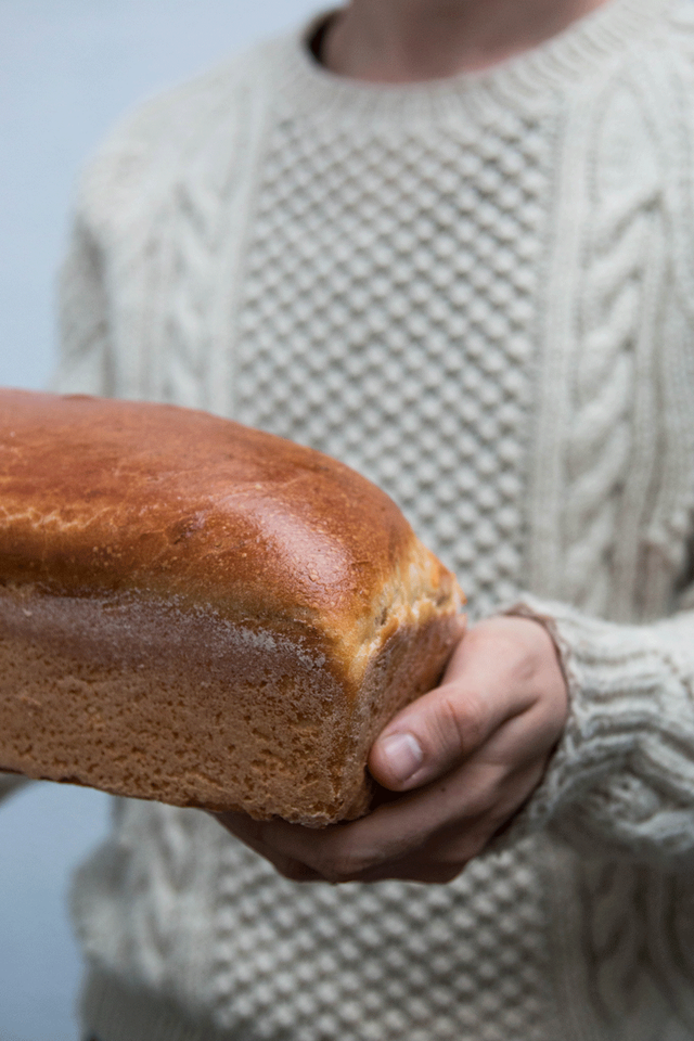 Bochník domácího toastového chleba se zlatohnědou kůrkou v rukou před bílým pleteným svetrem.