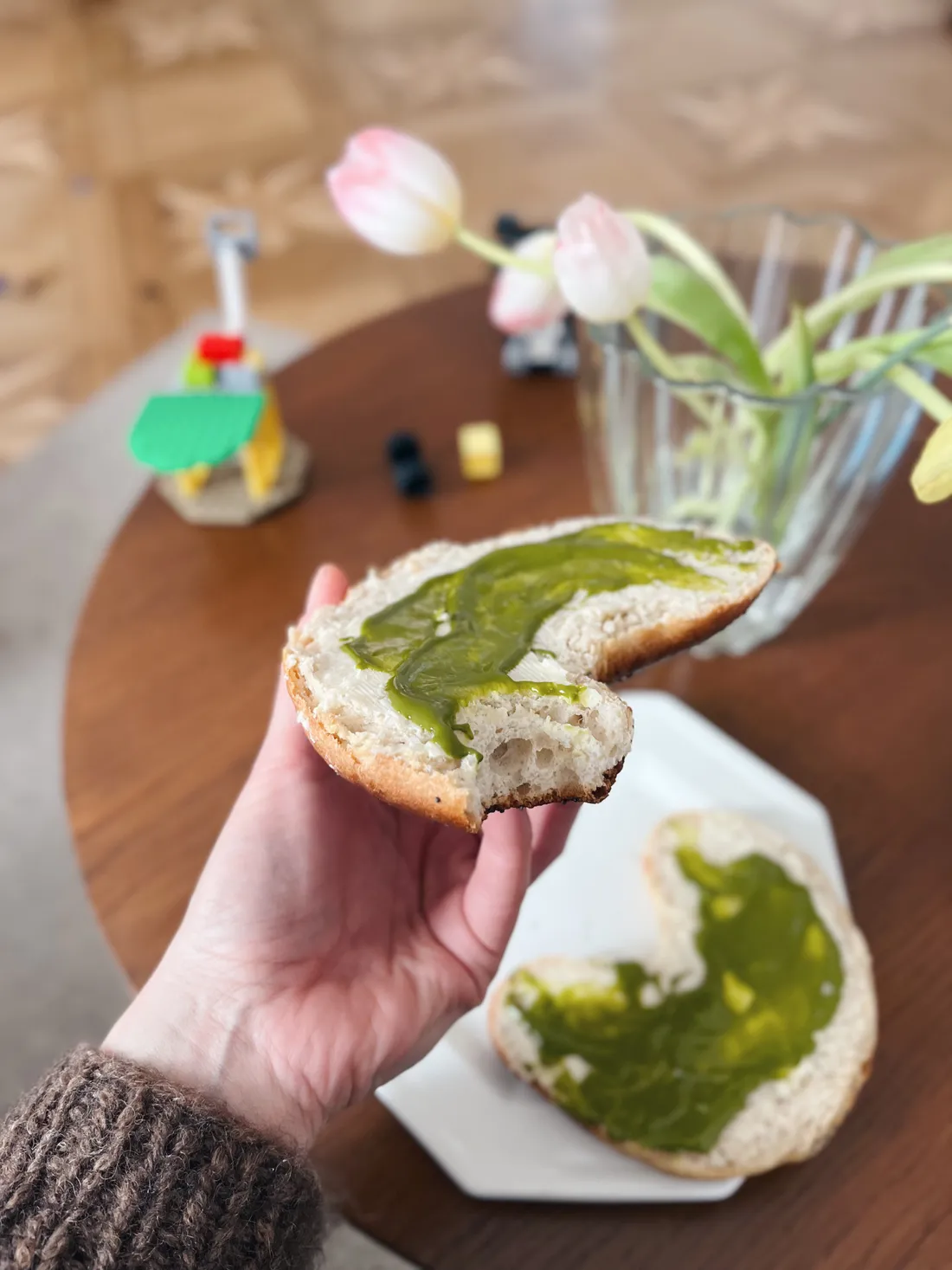 Ruka drží plátek toastu s hustou zelenou pistáciovou pomazánkou, vedle talíř s toustem a růžovými tulipány.