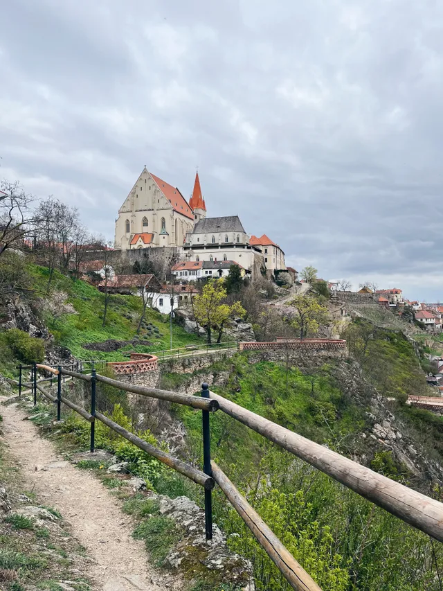 Pohled z lesní stezky s dřevěným zábradlím na gotický kostel s červenou střechou na skalnatém kopci.