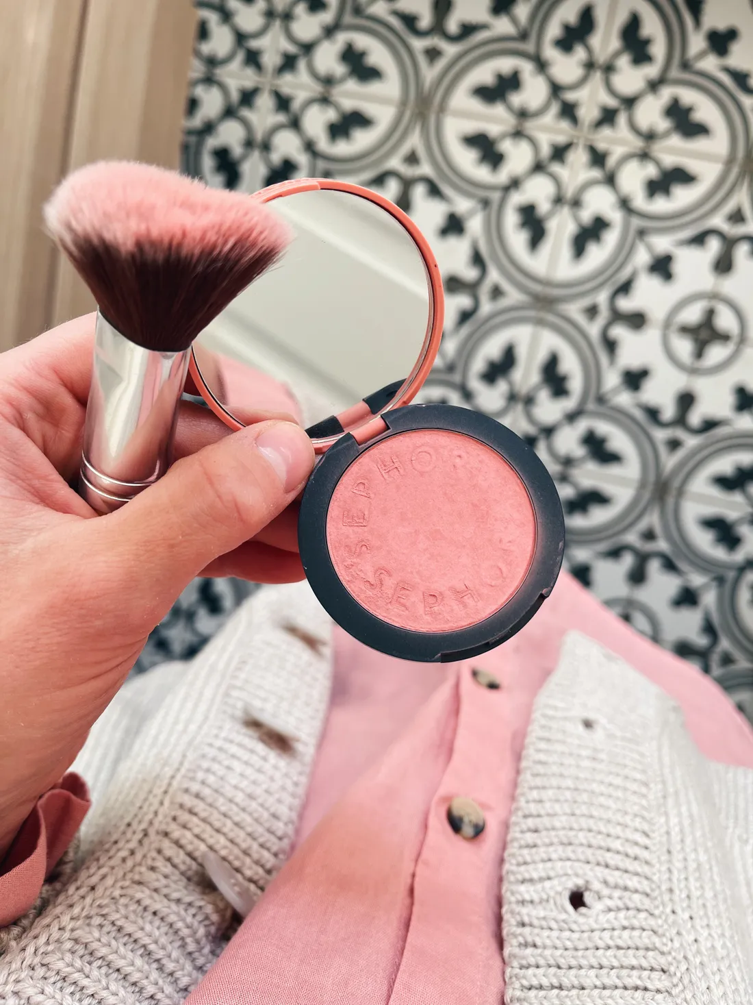 Ruka drží otevřenou růžovou tvářenku Sephora a kosmetický štětec před šedým svetrem a dlažbou.