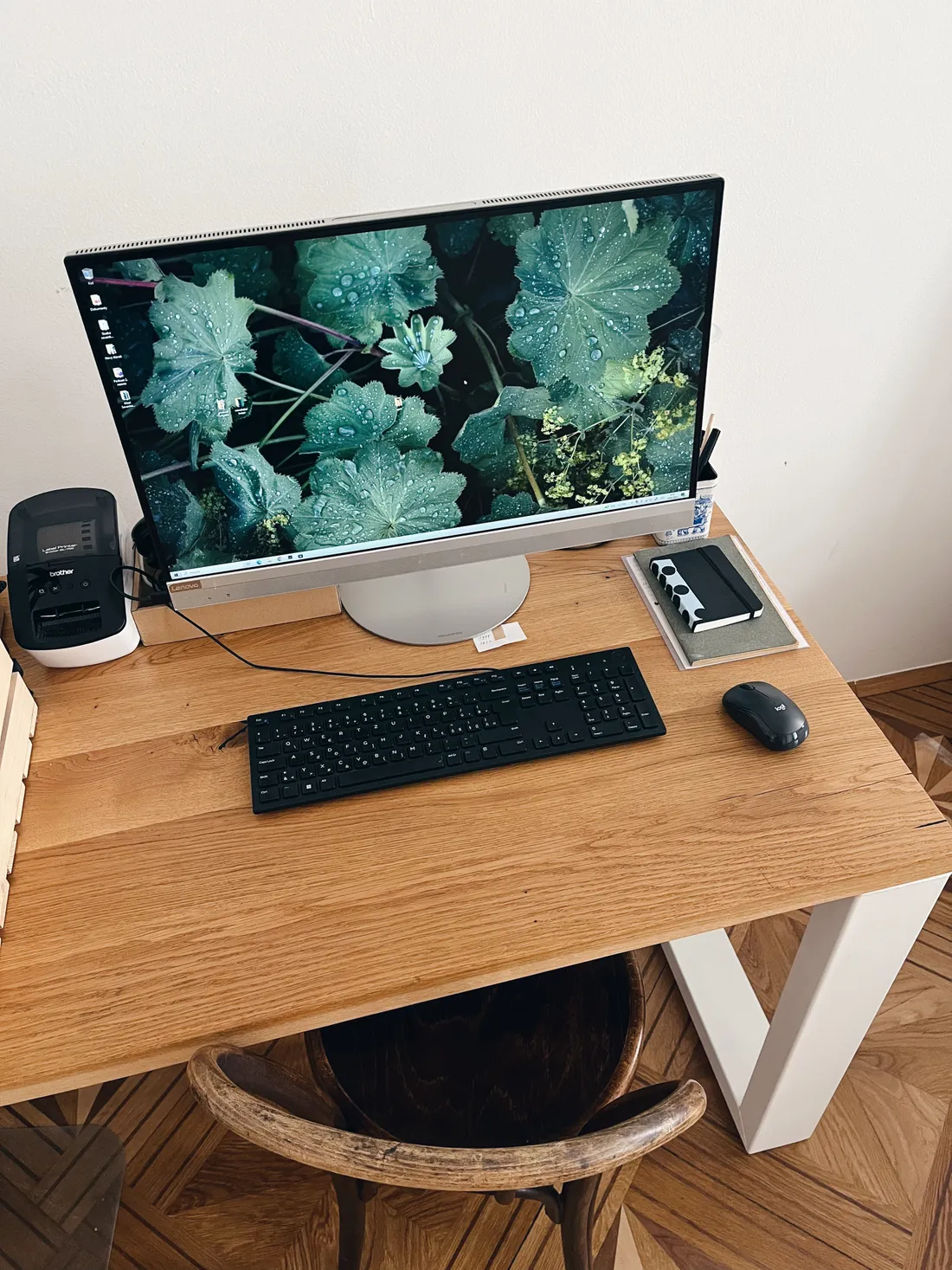 Pracovní stůl s monitorem s listy, černou klávesnicí, myší, tiskárnou štítků a zápisníky.