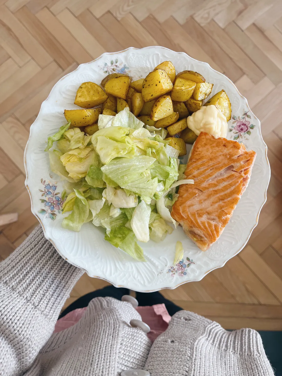Květovaný talíř s lososem, opečenými bramborami, ledovým salátem a kopečkem bílé omáčky v ruce nad parketami.