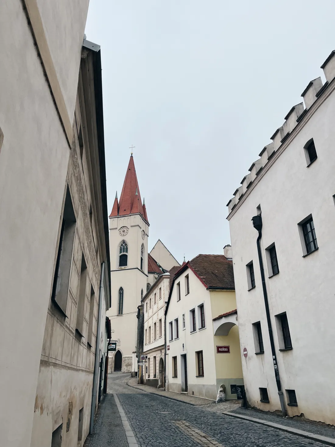 Pohled úzkou dlážděnou ulicí k bílé věži kostela s červenou střechou a hodinami pod šedou oblohou.