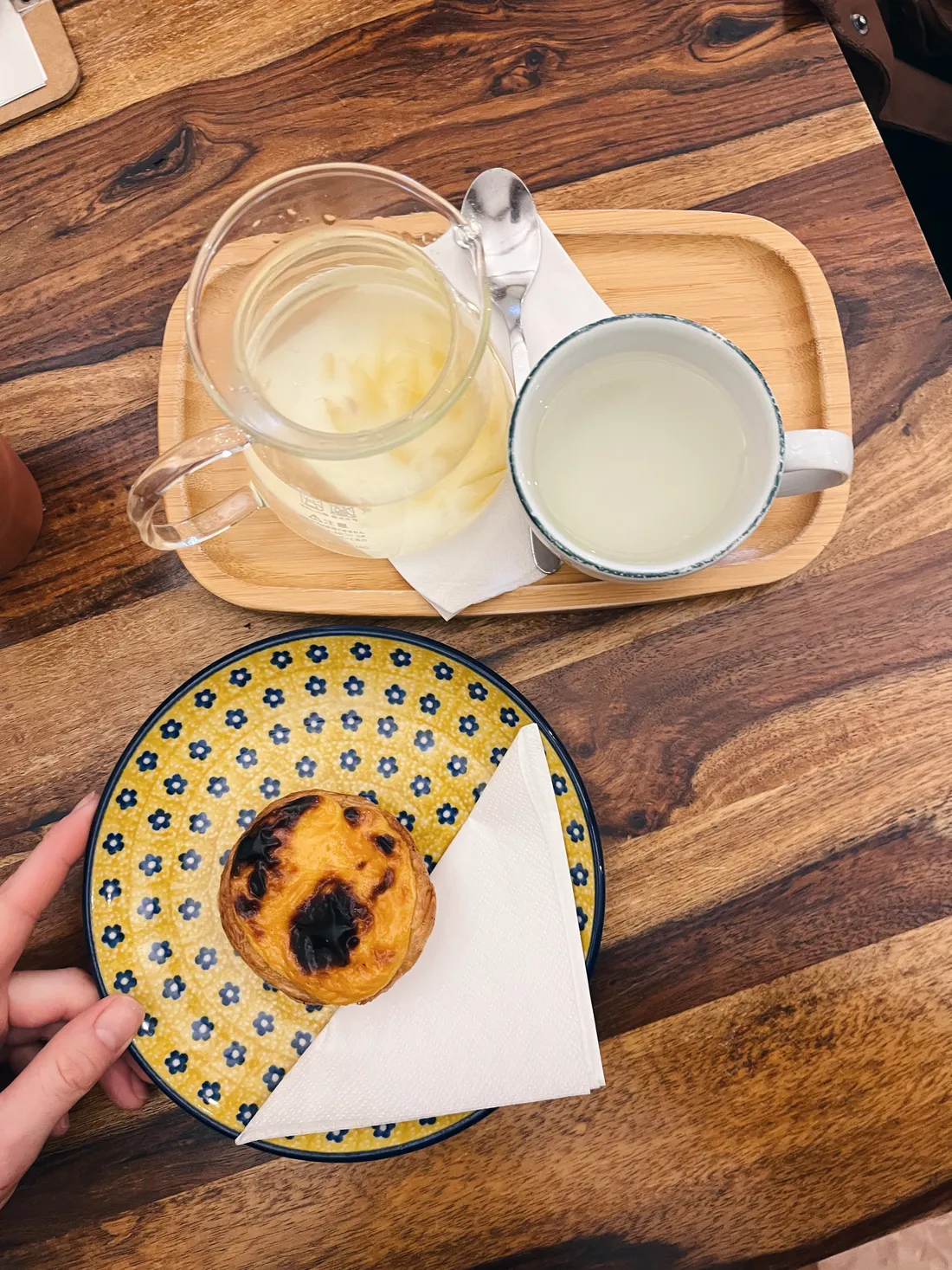 Konvička s čajem, hrnek a koláček Pastel de Nata na žlutém vzorovaném talíři na dřevěném stole.