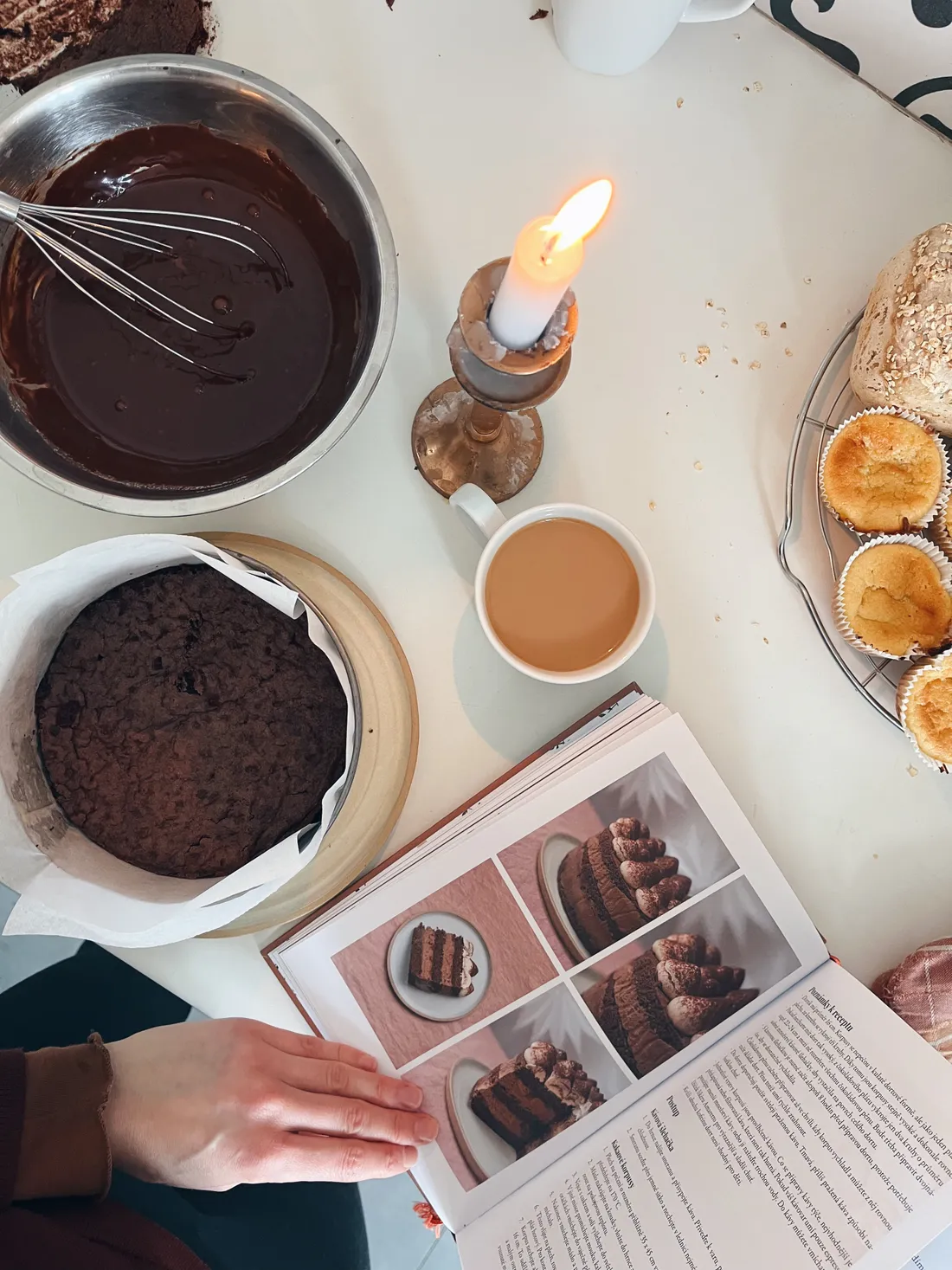 Příprava čokoládového dortu: báze, ganache, kuchařka, káva a svíčka na bílém stole.