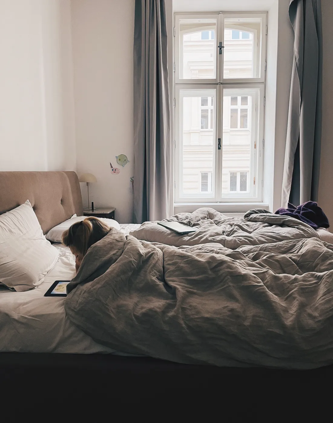 Osoba ležící v posteli pod šedou peřinou se dívá do tabletu, v pozadí okno se šedými závěsy.