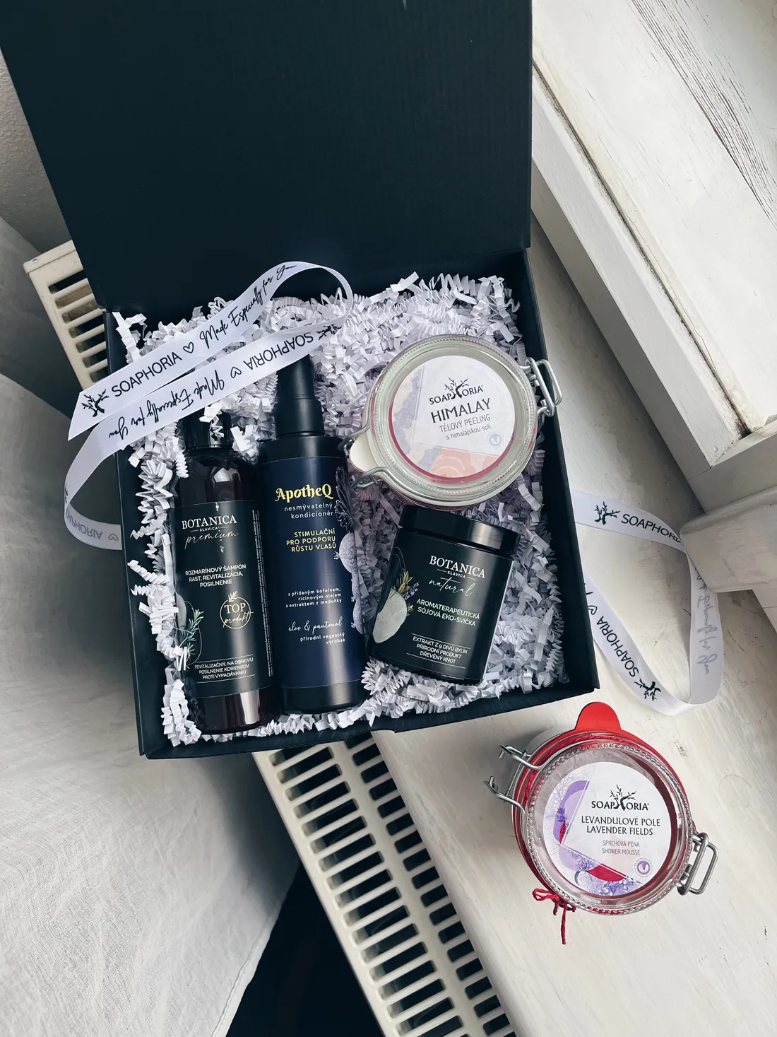 Otevřená černá dárková krabice s kosmetikou Soaphoria a ApotheQ na bílém parapetu vedle radiátoru.
