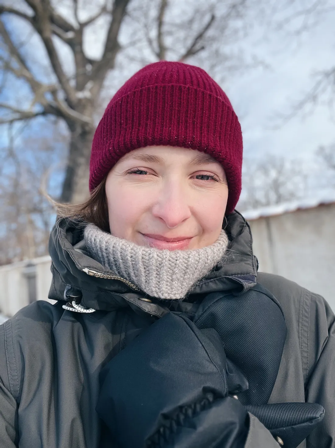 Usmívající se žena ve vínové čepici a šedém nákrčníku v zimní zahradě s holými stromy.