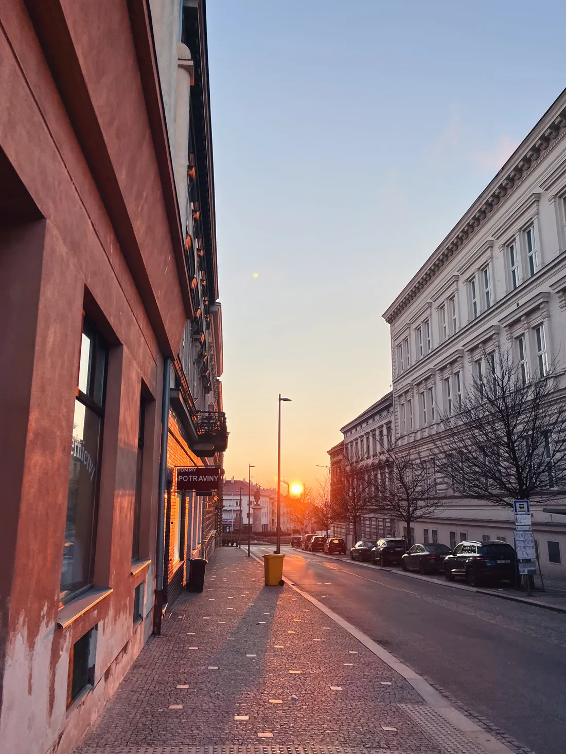Ulice při západu slunce mezi domy, v dálce oranžové slunce a cedule Potraviny.