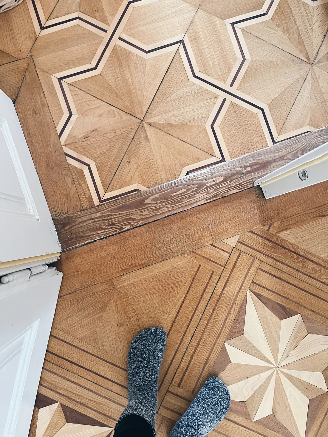 Dvě různé dřevěné parkety s hvězdovými vzory oddělené prahem, dole nohy v šedých ponožkách.