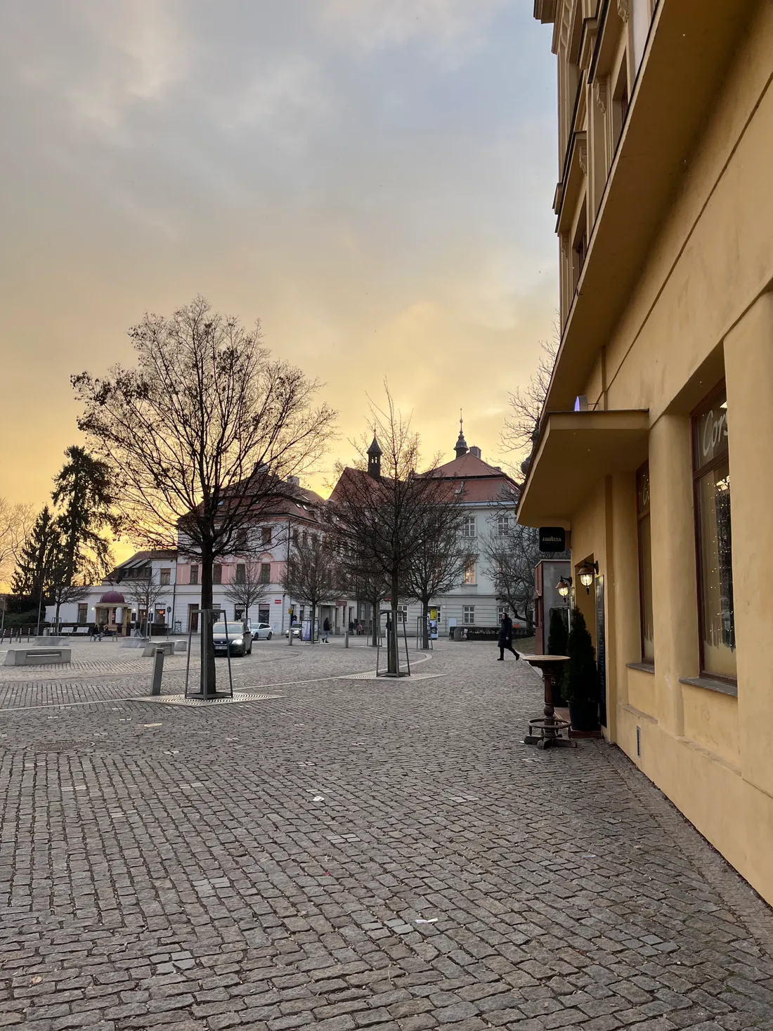 Dlážděné náměstí s holými stromy a historickými domy za zářivým západem slunce.