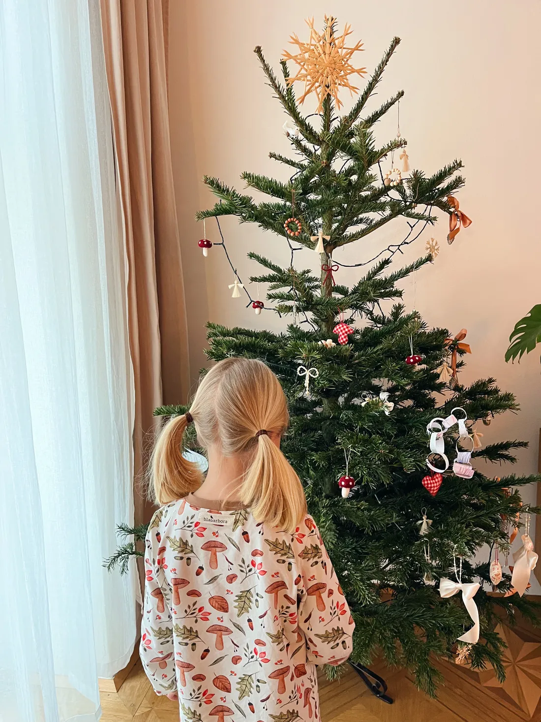 Dívka v houbovém tričku u vánočního stromku s červenými houbičkami a mašlemi.