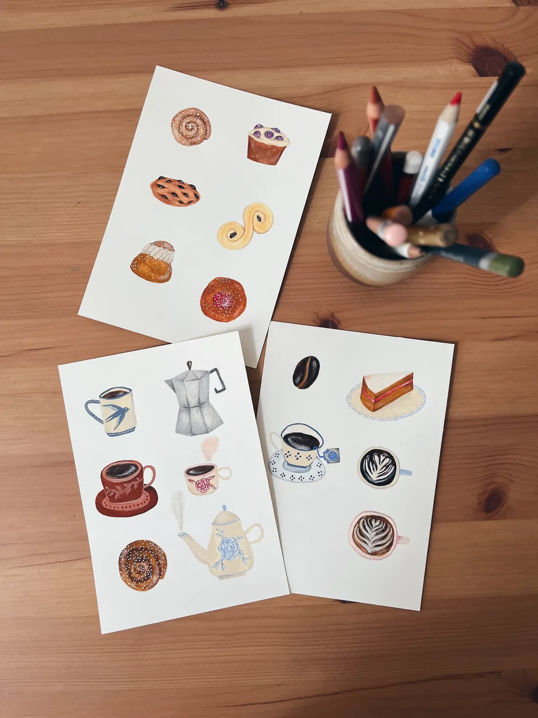 Tři karty s akvarelovými ilustracemi pečiva a kávy a kelímek pastelů na dřevěném stole.