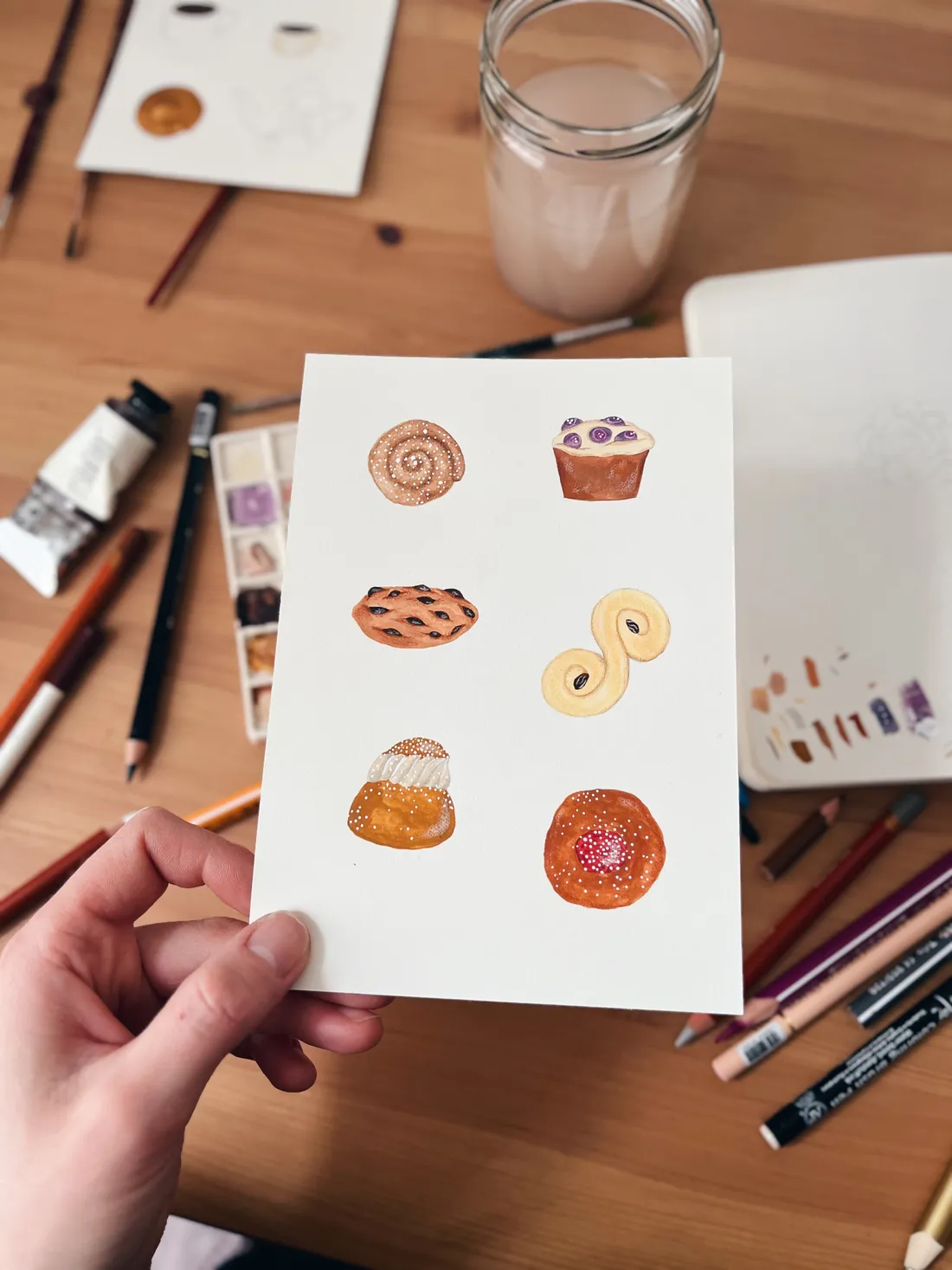 Ruka drží kartičku se šesti akvarelovými ilustracemi pečiva nad stolem s malířskými potřebami.