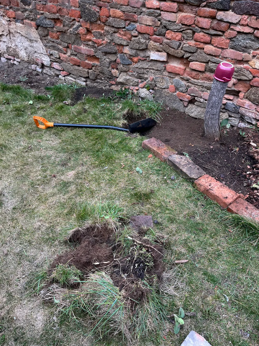 Zahrada s vykopanou dírou v trávě, rýč s oranžovou rukojetí a stará cihlová zeď v pozadí.