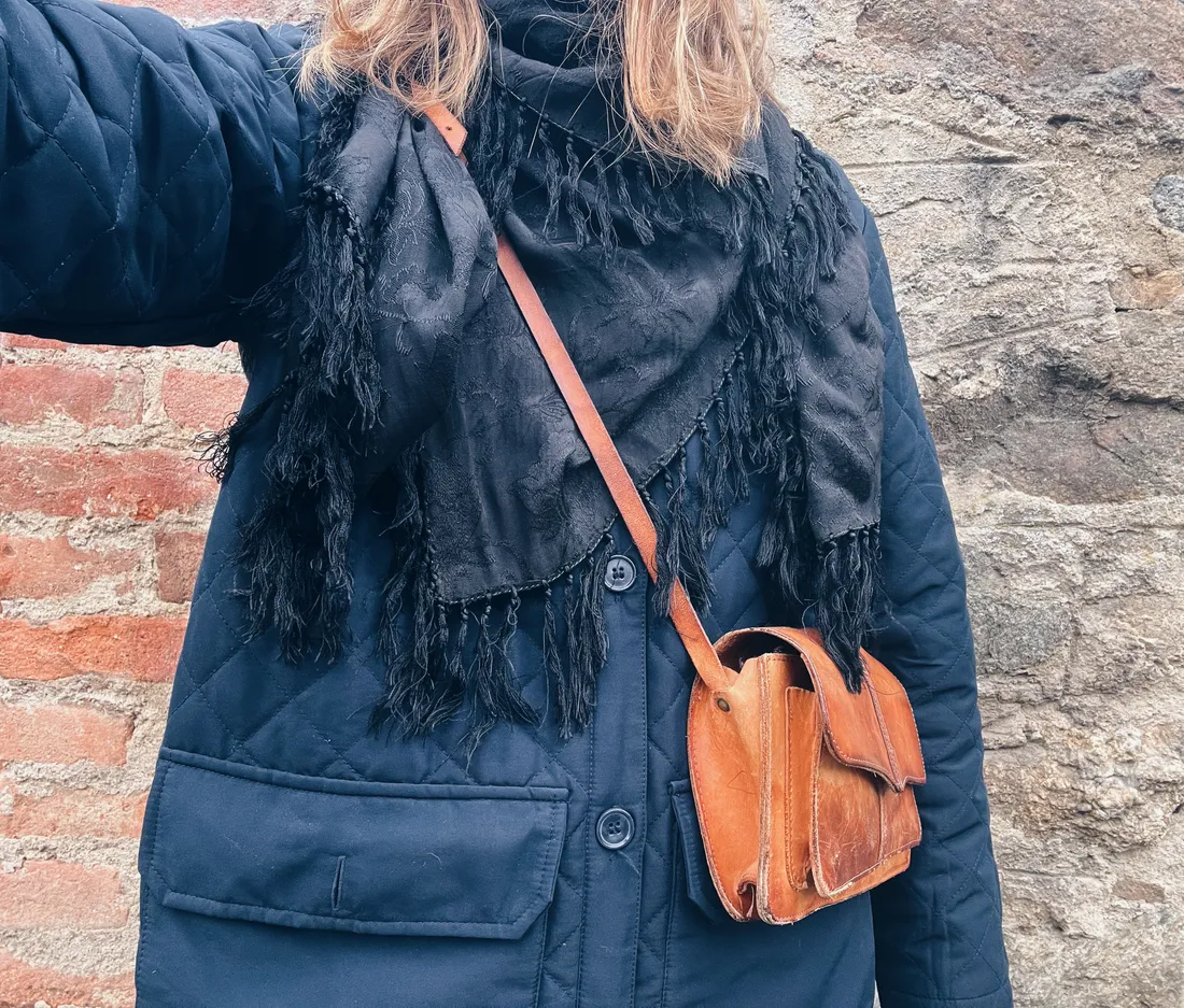 Tmavě modrá prošívaná bunda, černý šál s třásněmi a kožená crossbody taška u zdi.