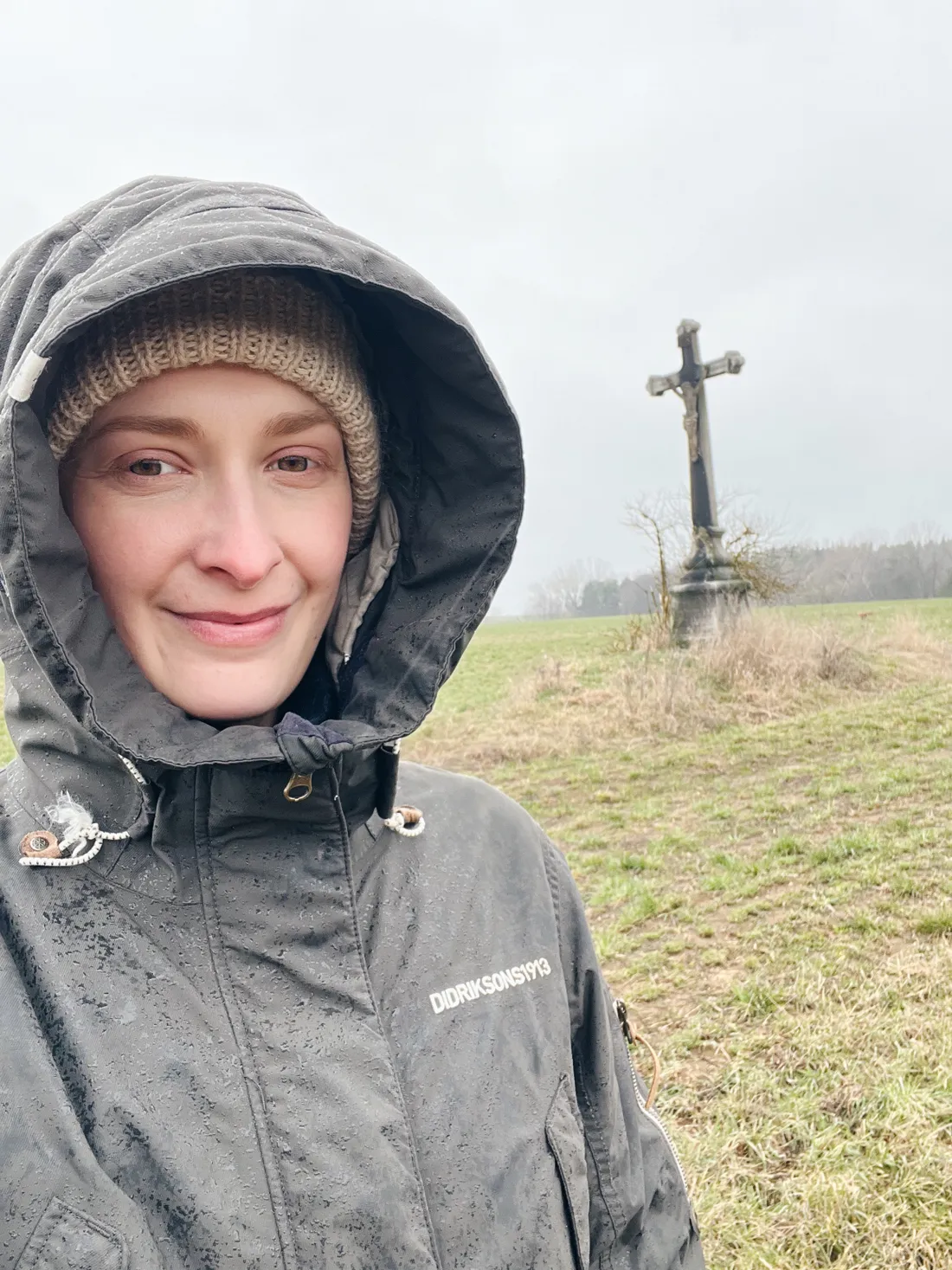 Usmívající se žena v kapuci s kapkami deště, v pozadí kamenná boží muka na louce.