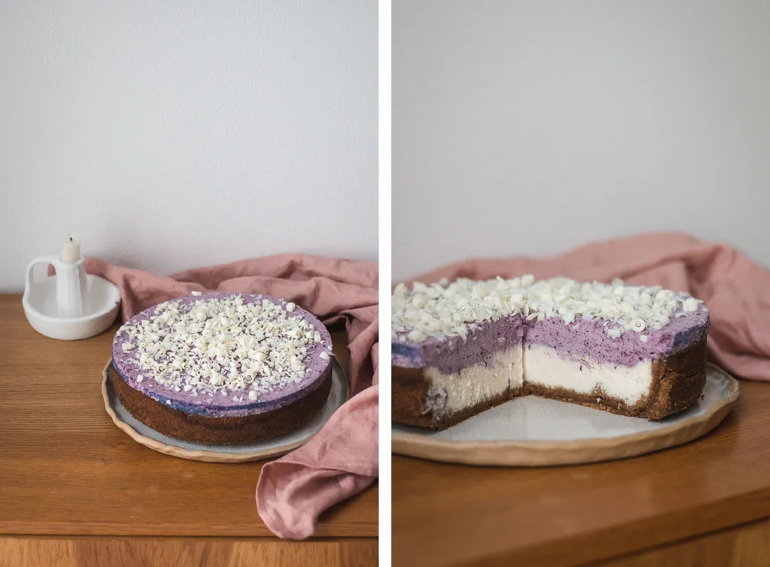 Borůvkový cheesecake s hoblinkami bílé čokolády na dřevěném stole – celý dort a řez se třemi vrstvami.